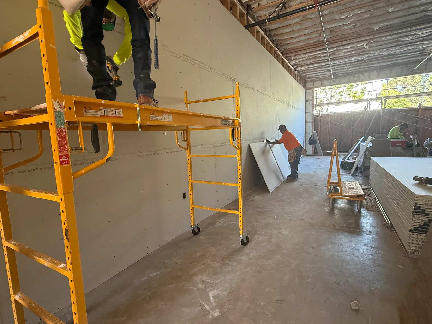 Drywall crew hanging board from rolling scaffold beside a stack of drywall sheets