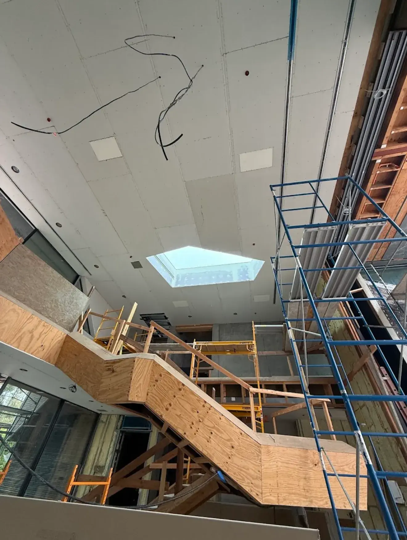 Drywall hung across a high stairwell ceiling around a skylight, with scaffolding and framed stairs below