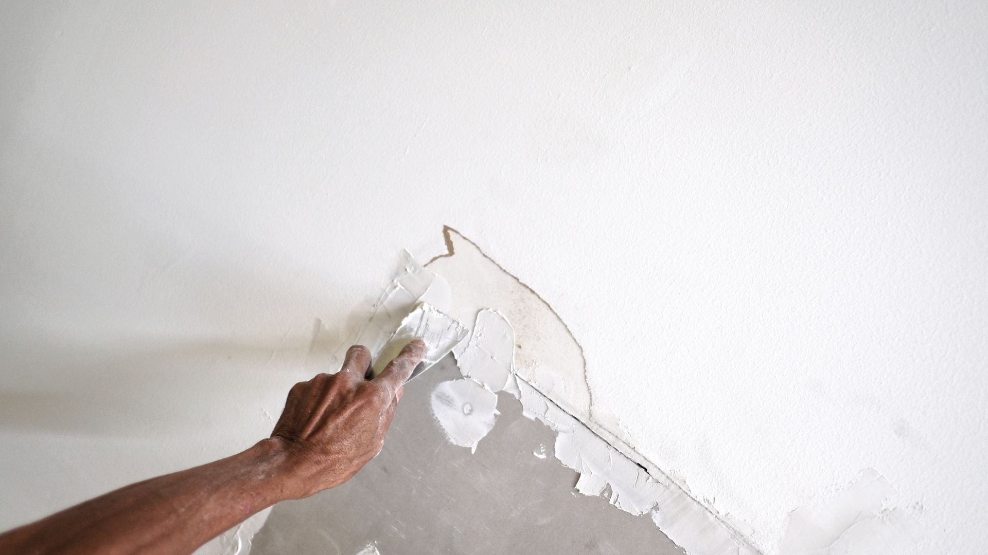 Hand peeling paint or wallpaper from white wall during renovation