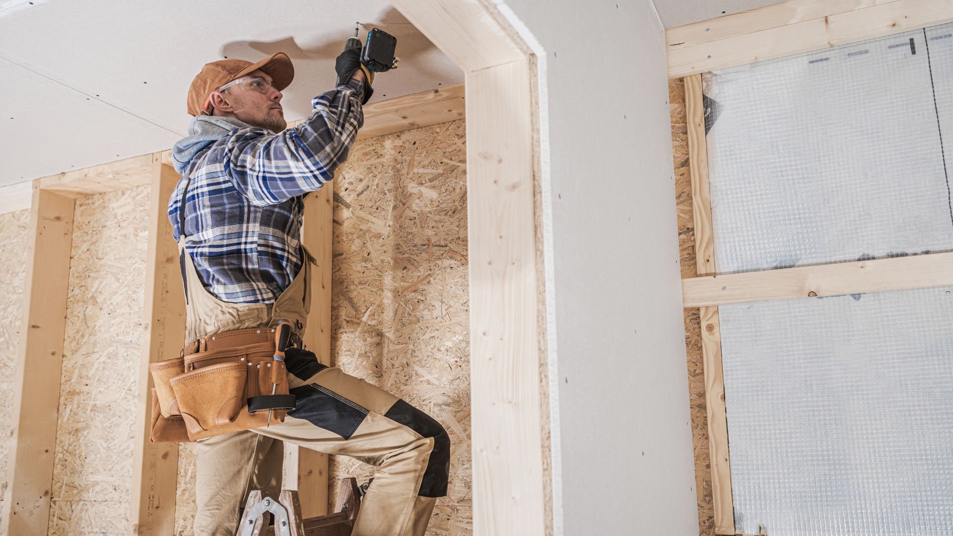 Construction worker installing drywall with power drill in wooden framed room